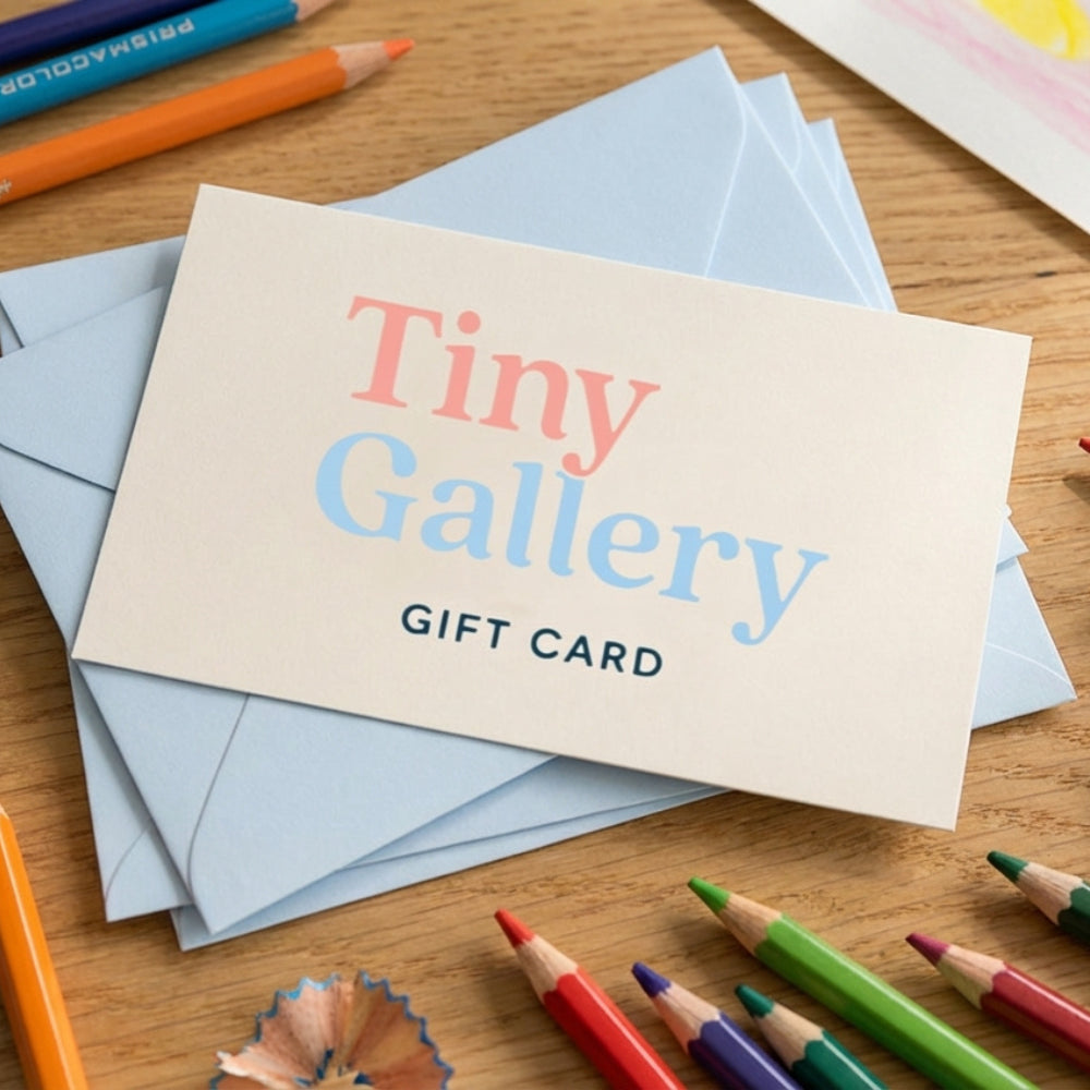 Vierkante 1:1 afbeelding van een Tiny Gallery gift card op een houten tafel. De cadeaubon ligt bovenop lichtblauwe enveloppen en wordt omringd door kleurpotloden en een geslepen potloodrestje. De setting heeft een warme, creatieve sfeer en benadrukt dat de gift card een perfect cadeau is voor ouders die kindertekeningen willen laten omzetten in een persoonlijk kunstboek.