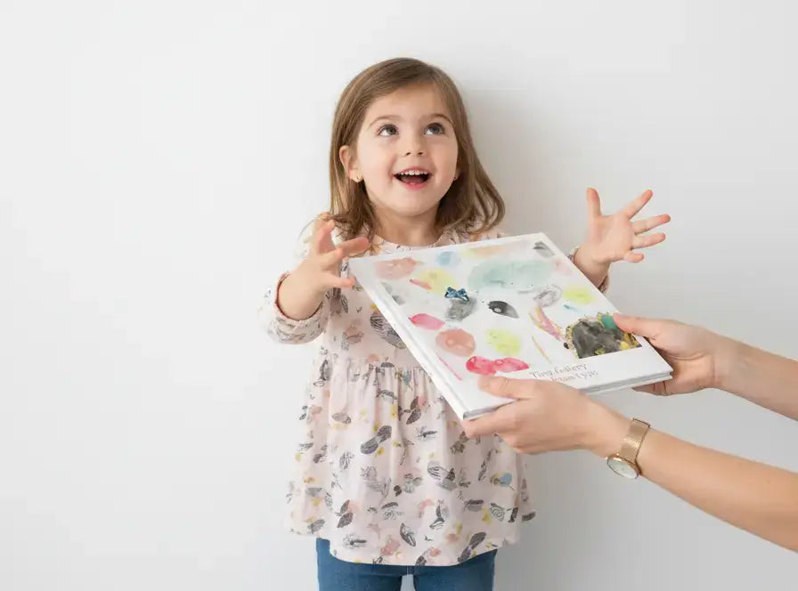 Een jong meisje kijkt blij en enthousiast terwijl ze een Tiny Gallery kunstboek aangereikt krijgt. Ze houdt haar armen open en kijkt omhoog, klaar om het boek vast te pakken.
