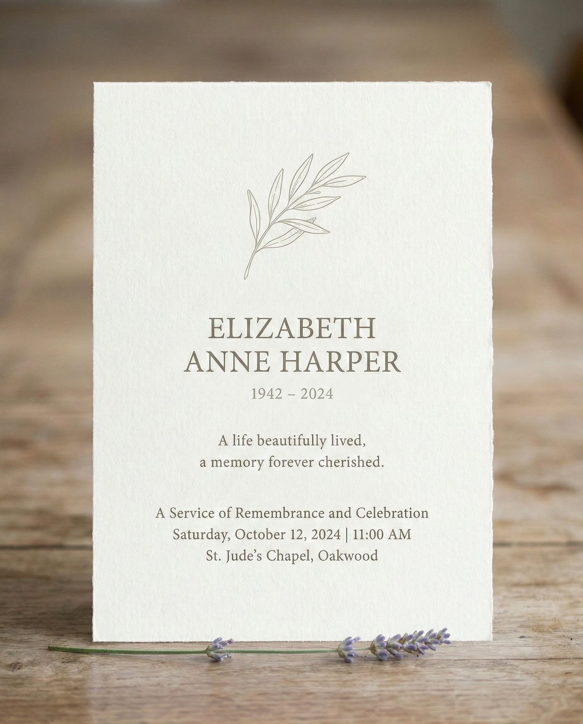 Crèmekleurige rouwkaart met een getekend olijftakje en de naam Elizabeth Anne Harper, 1942–2024, waarvan het ontwerp terugkomt in een Tiny Gallery rouwkaartenboek, op een houten tafel met lavendel