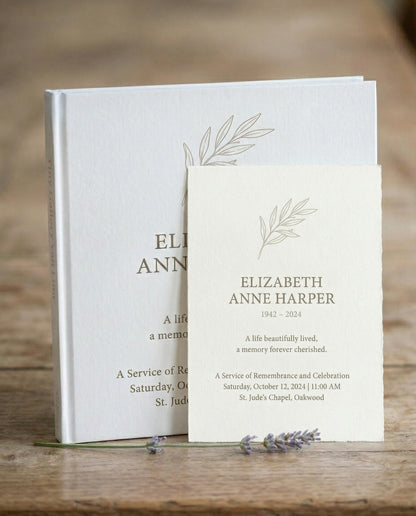 Wit Tiny Gallery kunstboek als herinneringsboek met een getekend olijftakje op de omslag, met ervoor de bijpassende rouwkaart van Elizabeth Anne Harper, op een houten tafel met lavendel