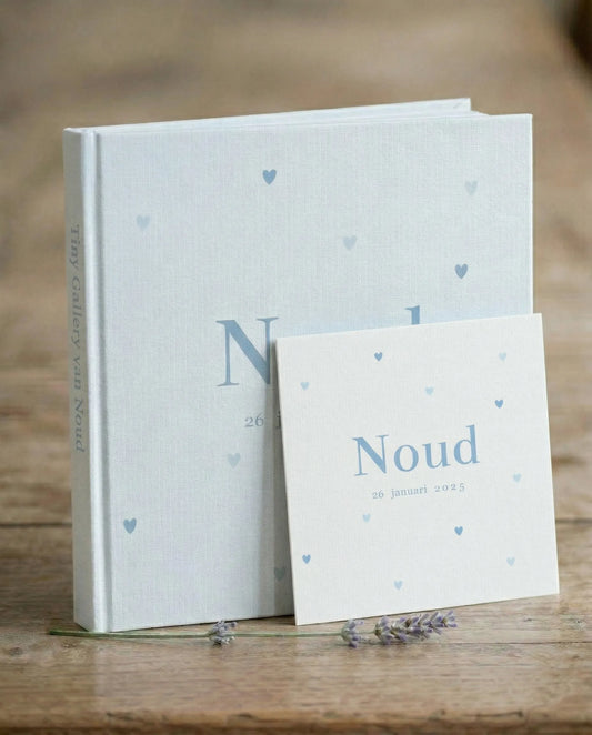 Lichtblauw linnen Tiny Gallery kunstboek ‘Tiny Gallery van Noud’ met hartjes op de omslag, met ervoor het bijpassende geboortekaartje met de naam Noud en 26 januari 2025, op een houten tafel met lavendel
