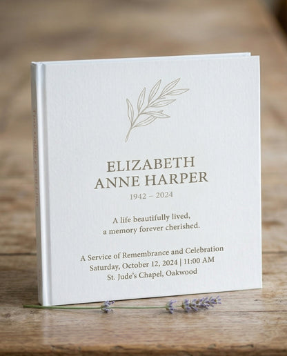Wit Tiny Gallery rouwkaartenboek met een getekend olijftakje en de naam Elizabeth Anne Harper, 1942–2024, op de omslag, staand op een houten tafel met lavendel