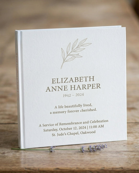 Wit Tiny Gallery rouwkaartenboek met een getekend olijftakje en de naam Elizabeth Anne Harper, 1942–2024, op de omslag, staand op een houten tafel met lavendel