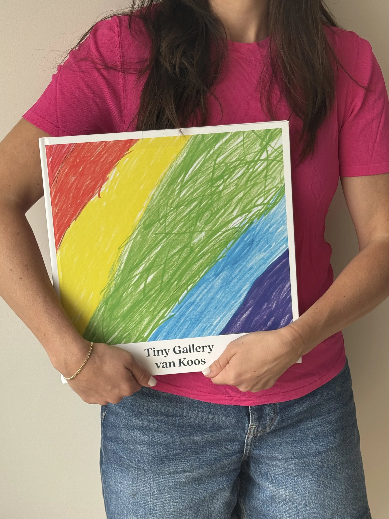 Vrouw in roze shirt en spijkerbroek houdt een groot Tiny Gallery-boek vast met een regenboogtekening op de kaft en de titel "Tiny Gallery van Koos".